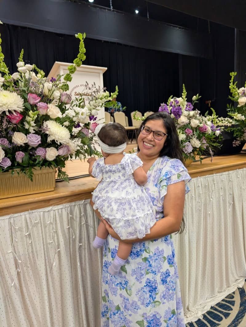 Zulamita holding her daughter, framed by floral arrangements at church